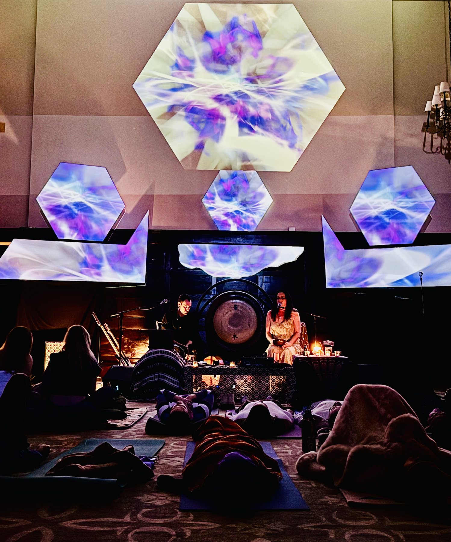 Purple and gold kaleidoscope projections glow in hexagons above Sariah and Taylor as the audience lies on mats