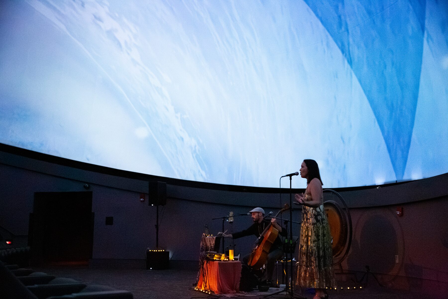 Sariah sings at the microphone as Taylor plays cello beneath a sweeping sky projection