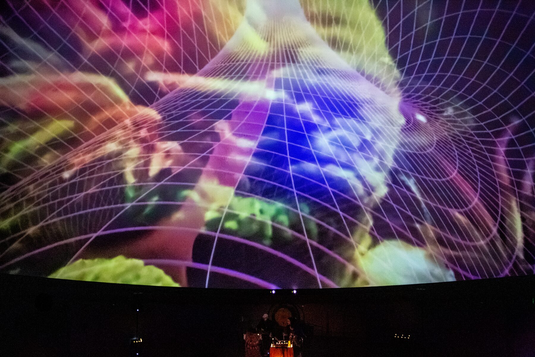 Neon green and violet light wraps a wireframe grid across the planetarium dome above the stage
