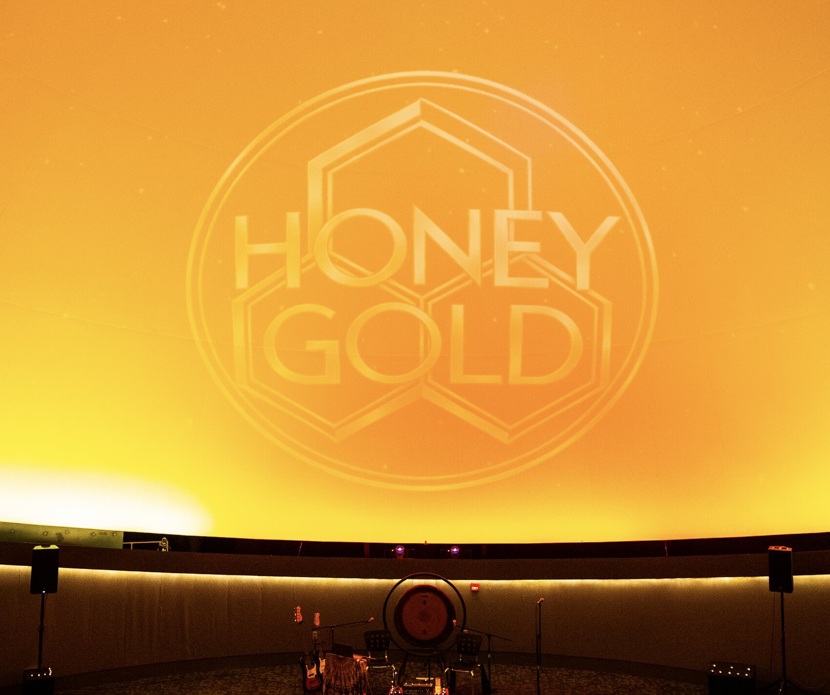 The Honey Gold logo glows across a golden dome above the gong and instruments waiting on stage