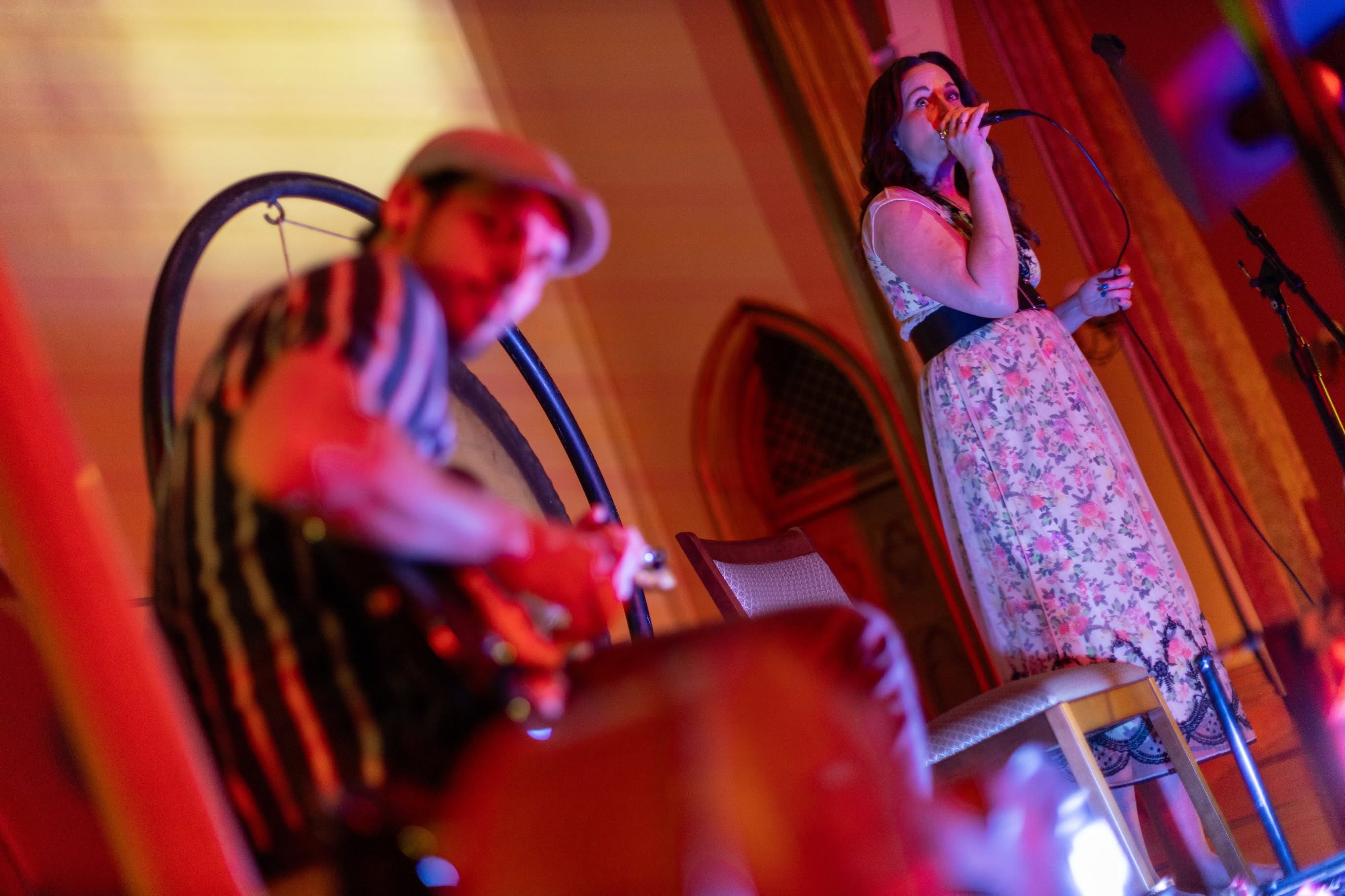 Sariah sings into the microphone in a floral dress as Taylor plays guitar in the warm red glow beside the gong