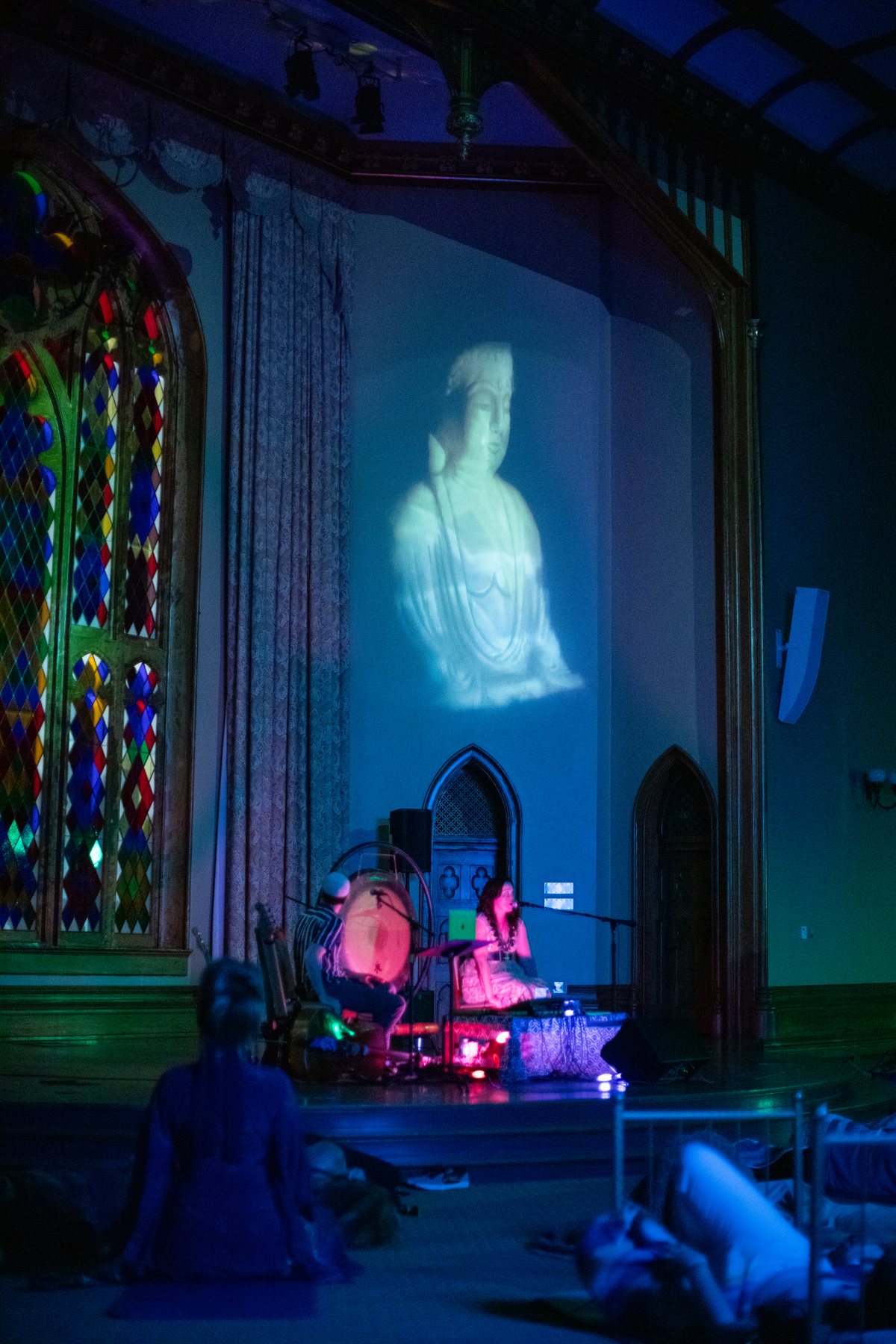 A glowing Buddha image projected above the stage as Sariah and Taylor perform for a resting audience
