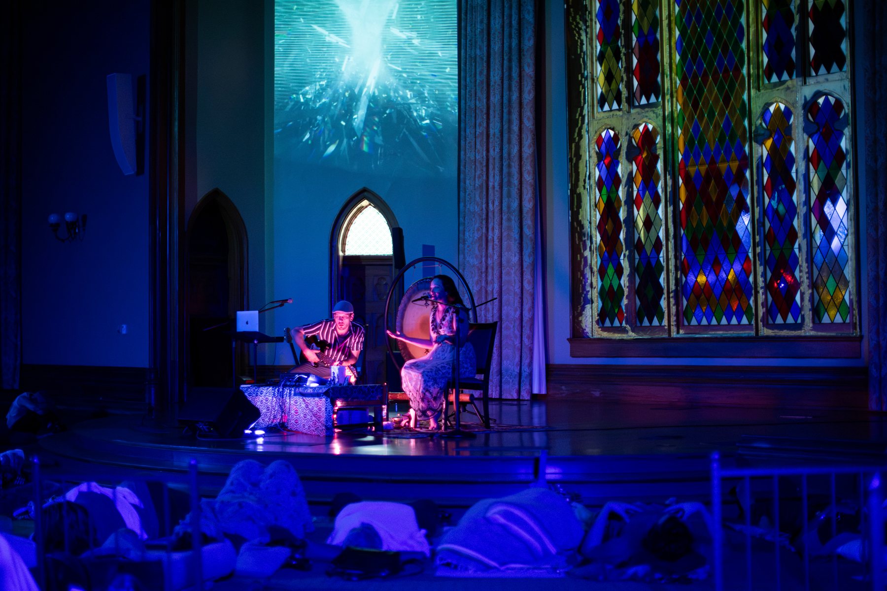 Sariah sings beside the gong and Taylor plays guitar beneath a crystalline projection and stained glass